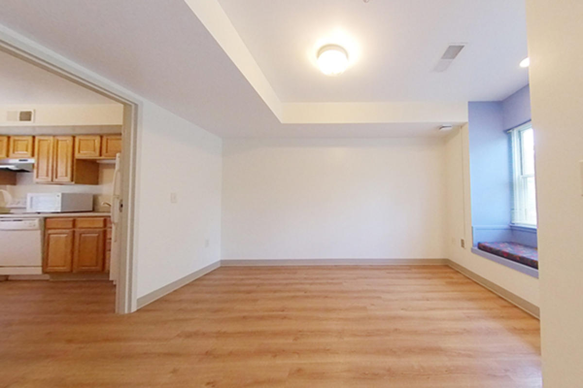 An empty living room with wooden floor, adjacent kitchen, and window.