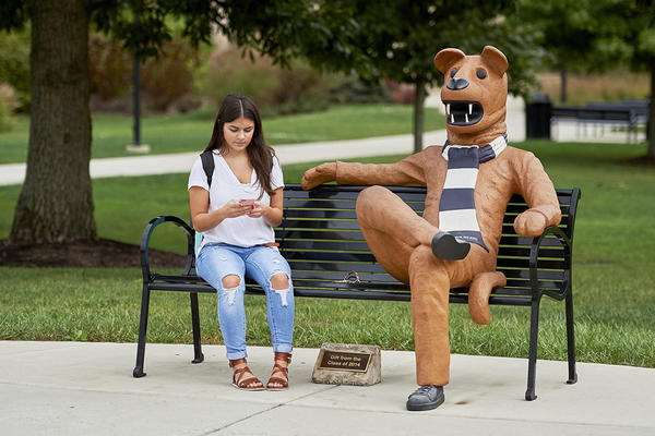 A girl checks her phone while seated on a bench next to the Nittany Lion mascot.