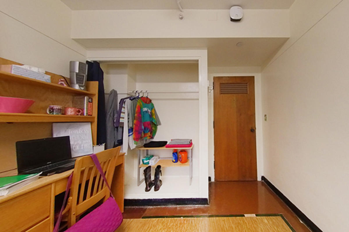 A tidy dorm room with wooden furniture and hanging clothes.