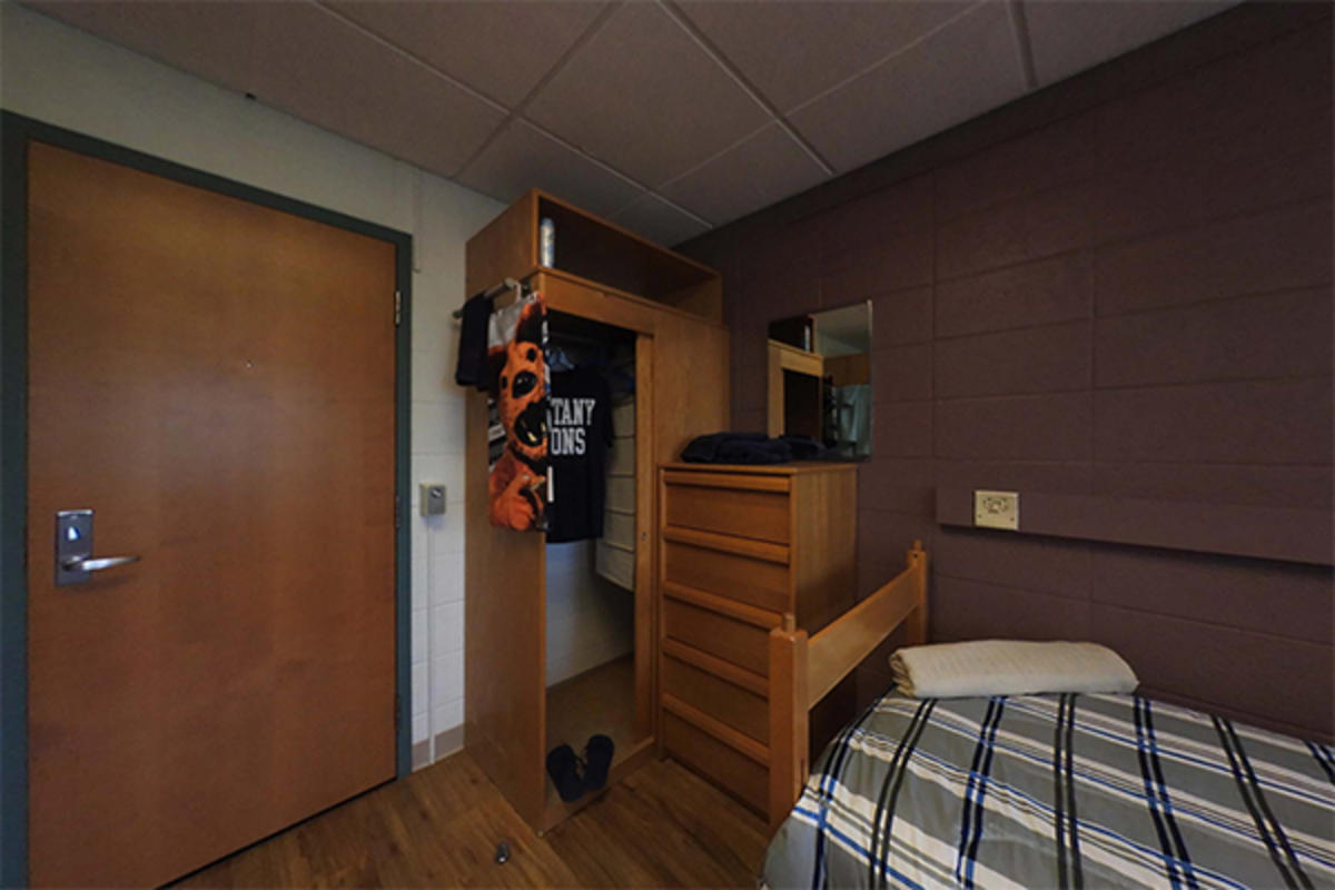 A tidy dorm room with a bed, wooden furniture, and hanging clothes.