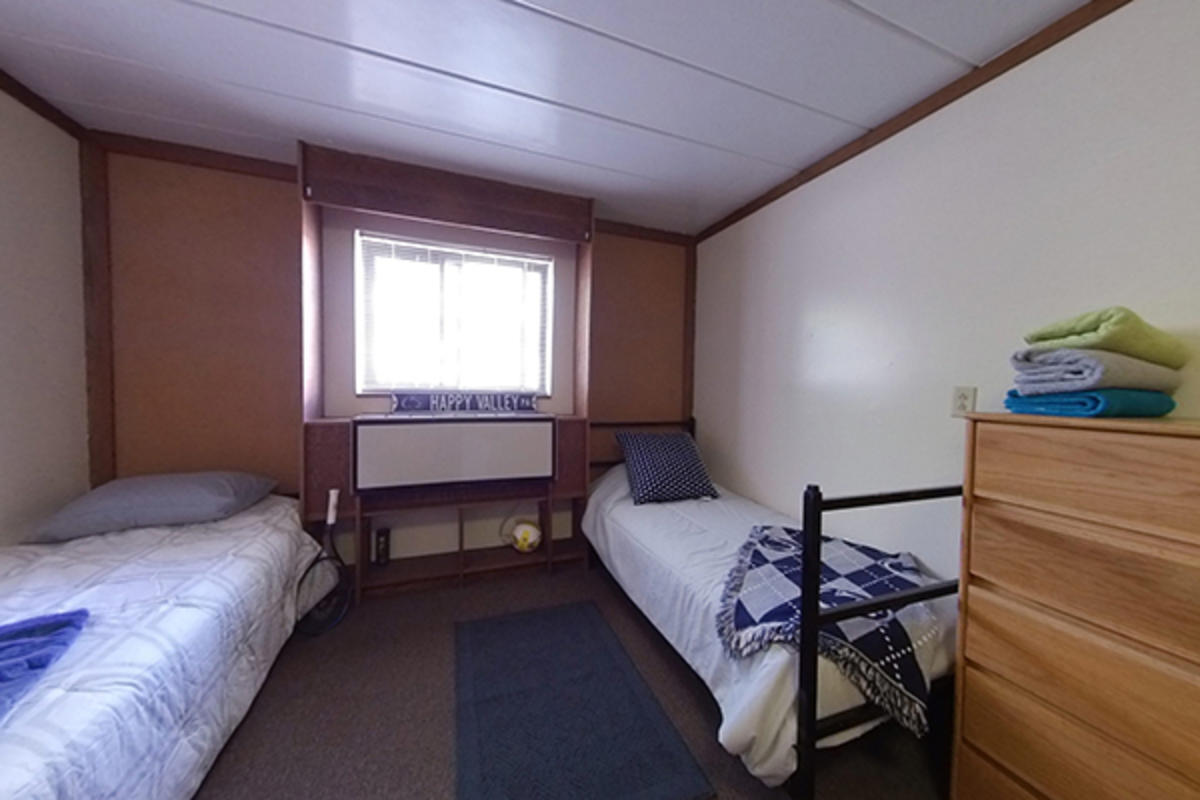 A tidy dorm room with two beds, a Nittany Lion blanket, and wooden furniture.