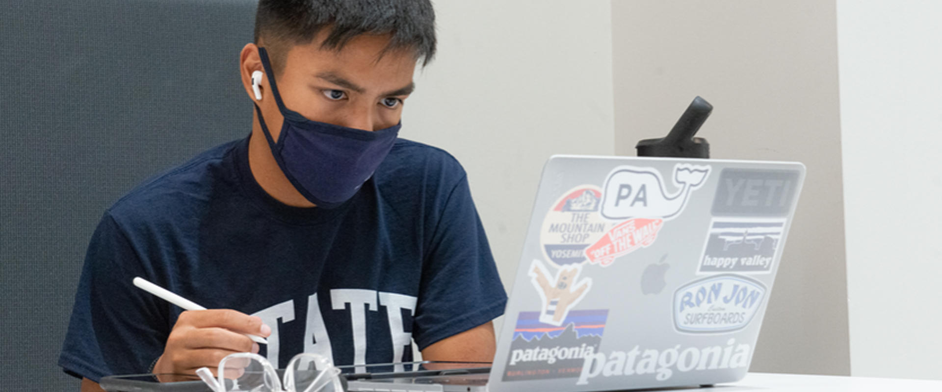 A student sits at a table with his laptop, wearing a mask 